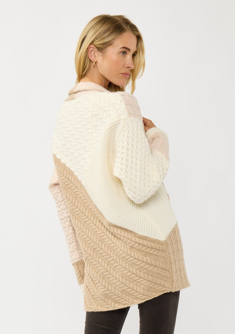 [Color: Natural/Tan] A back facing image of a blonde model wearing a pink and beige cable knit patchwork cardigan with an open front and cozy long sleeves. A cozy layering piece for the fall season. 