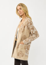 [Color: Sand] A side facing image of a blonde model wearing a brown floral print cardigan with a soft, fuzzy texture featuring an open front and patch pockets. 