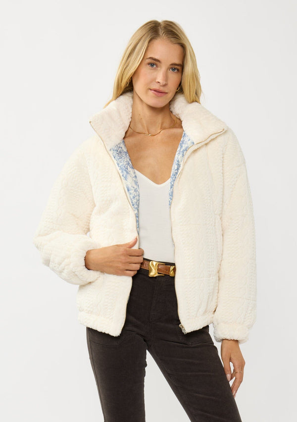 [Color: Cream] A front facing image of a blonde model wearing a plush cream patterned jacket with a zipper front, high collared neckline, contrast lining, and side pockets. A cozy fluffy jacket for the fall and winter season. 