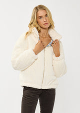 [Color: Cream] A front facing image of a blonde model wearing a plush cream patterned jacket with a zipper front, high collared neckline, contrast lining, and side pockets. A cozy fluffy jacket for the fall and winter season. 