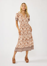 [Color: Natural/Brown] A front facing image of a blonde model wearing a bohemian floral border print mini dress with a split v-neckline, short sleeves, and an elastic waist. 
