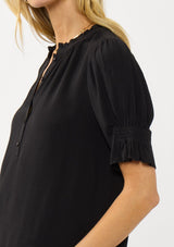 [Color: Black] A detail image of a blonde model wearing a casual black puff sleeve top with a ruffled neckline, button front, and a gathered sleeve detail. Effortless and versatile for easy layering or wearing on its own. 
