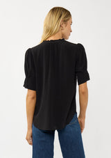 [Color: Black] A back facing image of a blonde model wearing a casual black puff sleeve top with a ruffled neckline, button front, and a gathered sleeve detail. Effortless and versatile for easy layering or wearing on its own. 