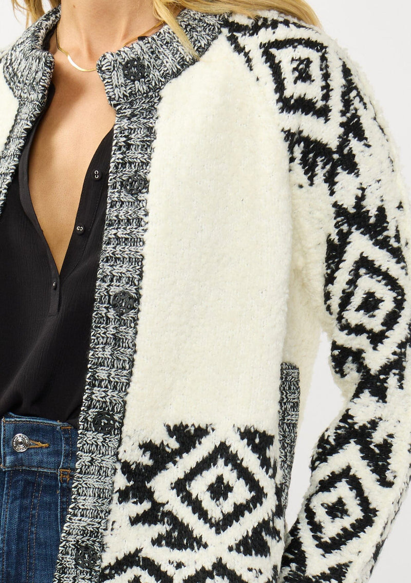 [Color: Winter White/Black] A detail image of a blonde model wearing a white and black tribal pattern sweater jacket with a popcorn texture, button front, and side pockets. A cozy fall sweater for day to night layering. 