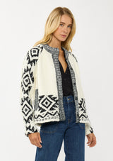 [Color: Winter White/Black] A side facing image of a blonde model wearing a white and black tribal pattern sweater jacket with a popcorn texture, button front, and side pockets. A cozy fall sweater for day to night layering. 