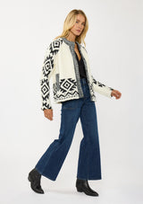 [Color: Winter White/Black] A side facing image of a blonde model wearing a white and black tribal pattern sweater jacket with a popcorn texture, button front, and side pockets. A cozy fall sweater for day to night layering. 