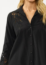 [Color: Black] A detail image of a blonde model wearing a black shirt with embroidered cutout details on the sleeves and shoulders. Featuring a classic collar neck, button front, and button cuff closure. An elevated fall shirt. 