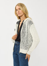 [Color: Cream/Black] A side facing image of a blonde model wearing a color block popcorn sweater jacket. A cozy textured sweater with a zipper front, hoodie, and long sleeves. 