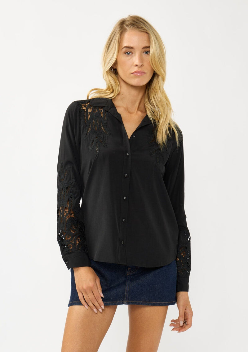 [Color: Black] A front facing image of a blonde model wearing a black shirt with embroidered cutout details on the sleeves and shoulders. Featuring a classic collar neck, button front, and button cuff closure. An elevated fall shirt. 