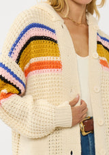 [Color: Natural/Mustard] A detail image of a blonde model wearing a cream knit cardigan with a colorful striped pattern. Laid back with a hoodie, patch pockets, and button front. A cool girl fall essential!