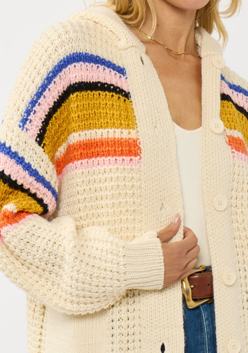 [Color: Natural/Mustard] A detail image of a blonde model wearing a cream knit cardigan with a colorful striped pattern. Laid back with a hoodie, patch pockets, and button front. A cool girl fall essential!