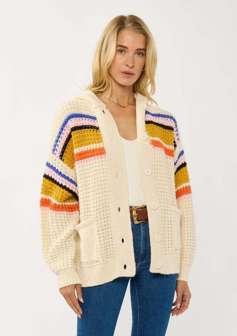 [Color: Natural/Mustard] A front facing image of a blonde model wearing a cream knit cardigan with a colorful striped pattern. Laid back with a hoodie, patch pockets, and button front. A cool girl fall essential!