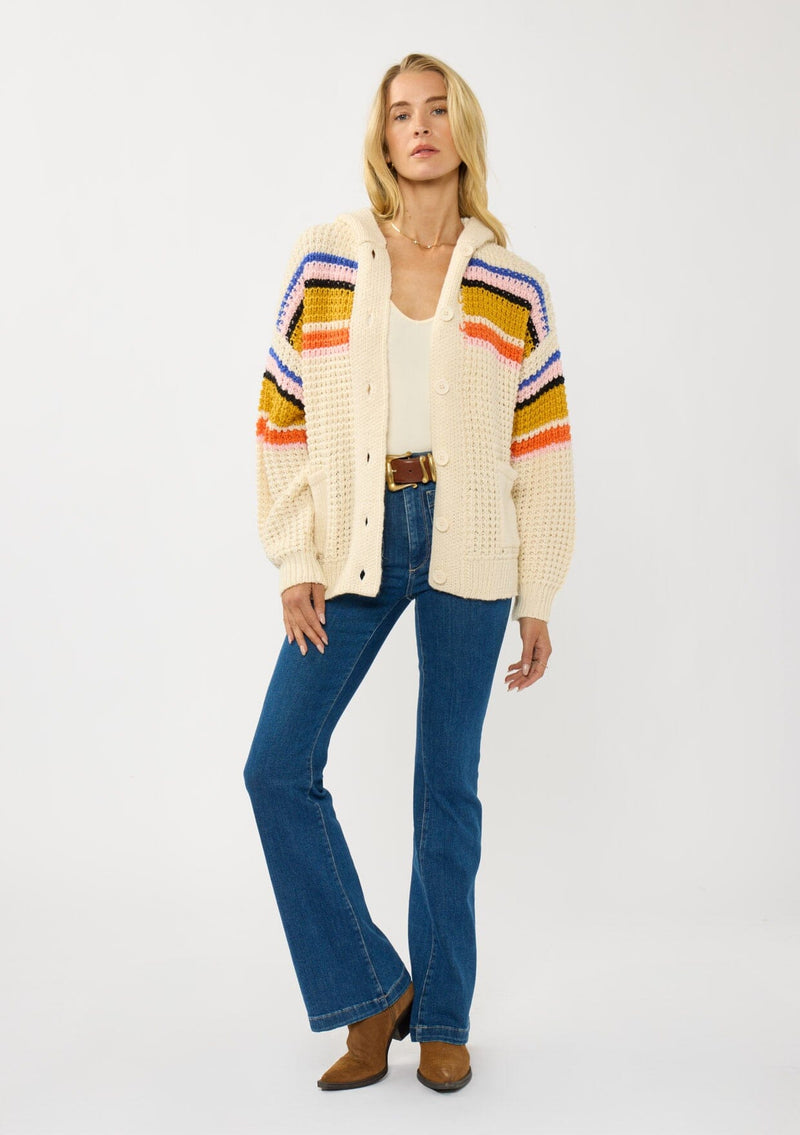 [Color: Natural/Mustard] A front facing image of a blonde model wearing a cream knit cardigan with a colorful striped pattern. Laid back with a hoodie, patch pockets, and button front. A cool girl fall essential!