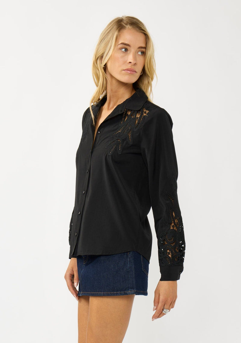 [Color: Black] A side facing image of a blonde model wearing a black shirt with embroidered cutout details on the sleeves and shoulders. Featuring a classic collar neck, button front, and button cuff closure. An elevated fall shirt. 