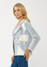 [Color: White/Dusty Blue] A side facing image of a blonde model wearing a blue striped sweater jacket with white geometric accents. Featuring a notched lapel, double-breasted front, snap buttons, and side pockets. A cozy and chic fall sweater for the office and casual outings. 