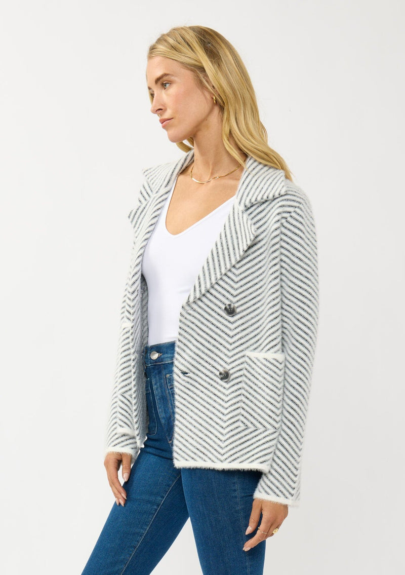 [Color: Black/White] A side facing image of a blonde model wearing a chevron stripe sweater jacket with a soft, fuzzy texture. Featuring a notched collar, double-breasted front, and patch pockets. Cozy and chic for cold weather styling. 