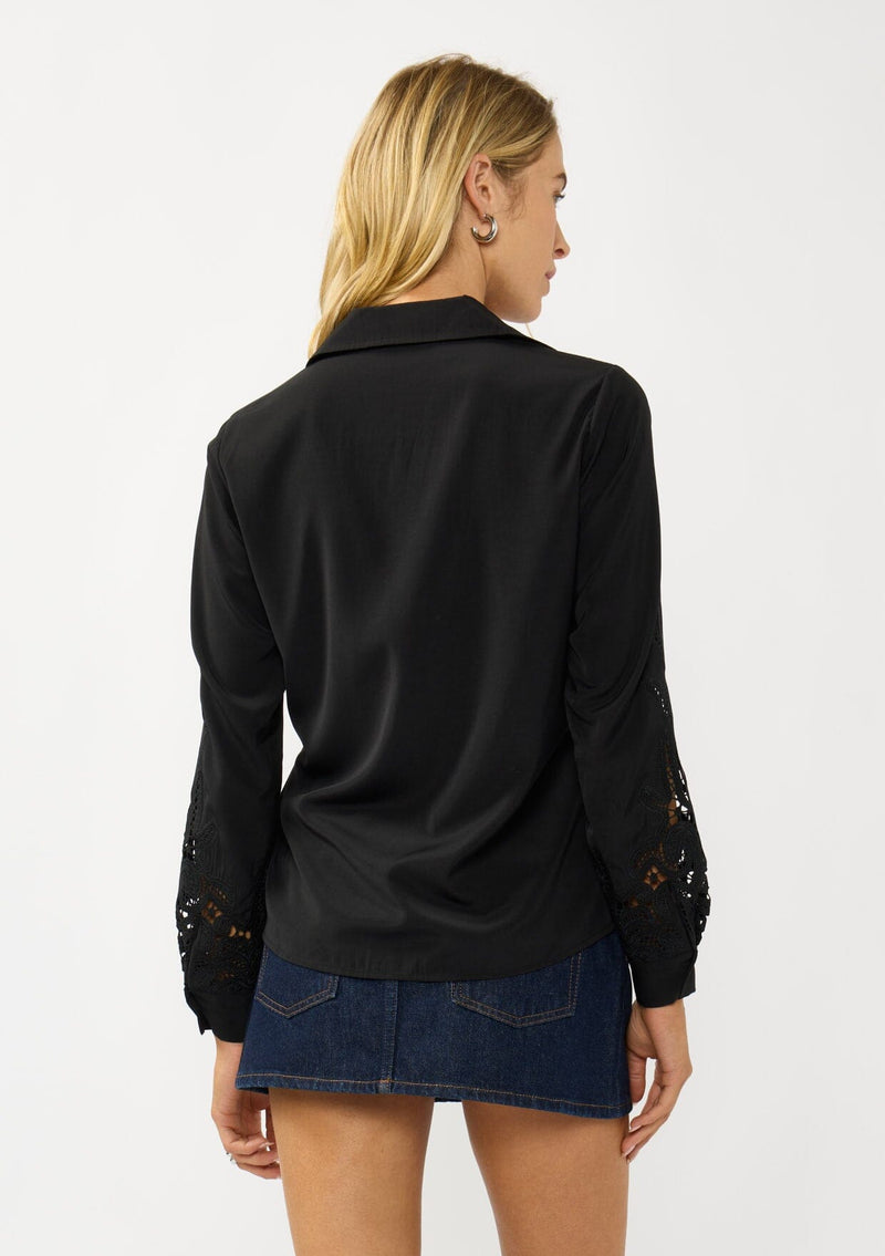 [Color: Black] A back facing image of a blonde model wearing a black shirt with embroidered cutout details on the sleeves and shoulders. Featuring a classic collar neck, button front, and button cuff closure. An elevated fall shirt. 