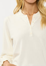 [Color: Cream] A detail image of a blonde model wearing a cozy sweater top with a button front, long sleeves, and ruffle trim details. Casual and chic for the fall season.