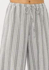 [Color: Grey/White] A detail image of a blonde model wearing a grey striped bottom crafted in airy cotton with an elastic drawstring waist and wide legs. Effortless and breezy for vacation and resort styling. 