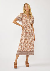 [Color: Natural/Brown] A front facing image of a blonde model wearing a bohemian floral border print mini dress with a split v-neckline, short sleeves, and an elastic waist. 