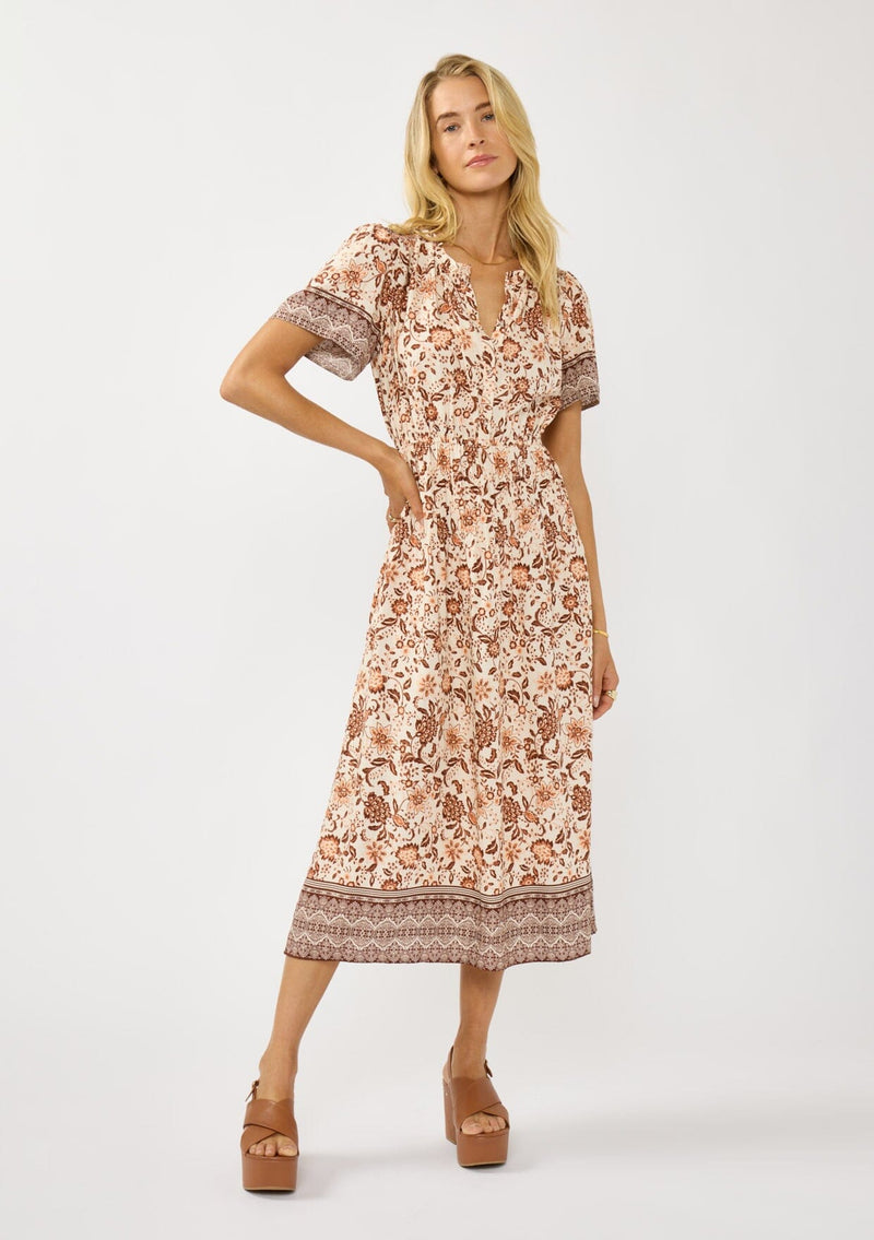 [Color: Natural/Brown] A front facing image of a blonde model wearing a bohemian floral border print mini dress with a split v-neckline, short sleeves, and an elastic waist. 