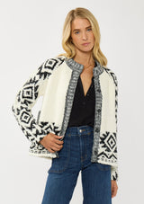 [Color: Winter White/Black] A front facing image of a blonde model wearing a white and black tribal pattern sweater jacket with a popcorn texture, button front, and side pockets. A cozy fall sweater for day to night layering. 