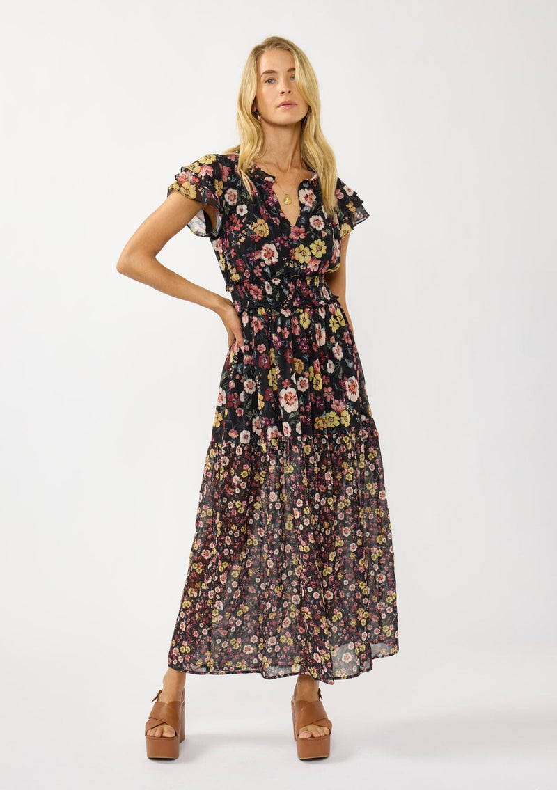 [Color: Black/Rust] A front facing image of a blonde model wearing a black floral maxi dress with short flutter sleeves, a split v-neckline, a smocked waist, and side slit. Adorned with ruffle trims, this romantic fall maxi dress is perfect for weddings, holidays, and other special occasions. 