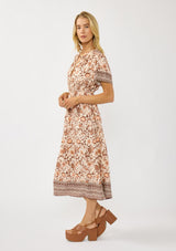 [Color: Natural/Brown] A side facing image of a blonde model wearing a bohemian floral border print mini dress with a split v-neckline, short sleeves, and an elastic waist. 
