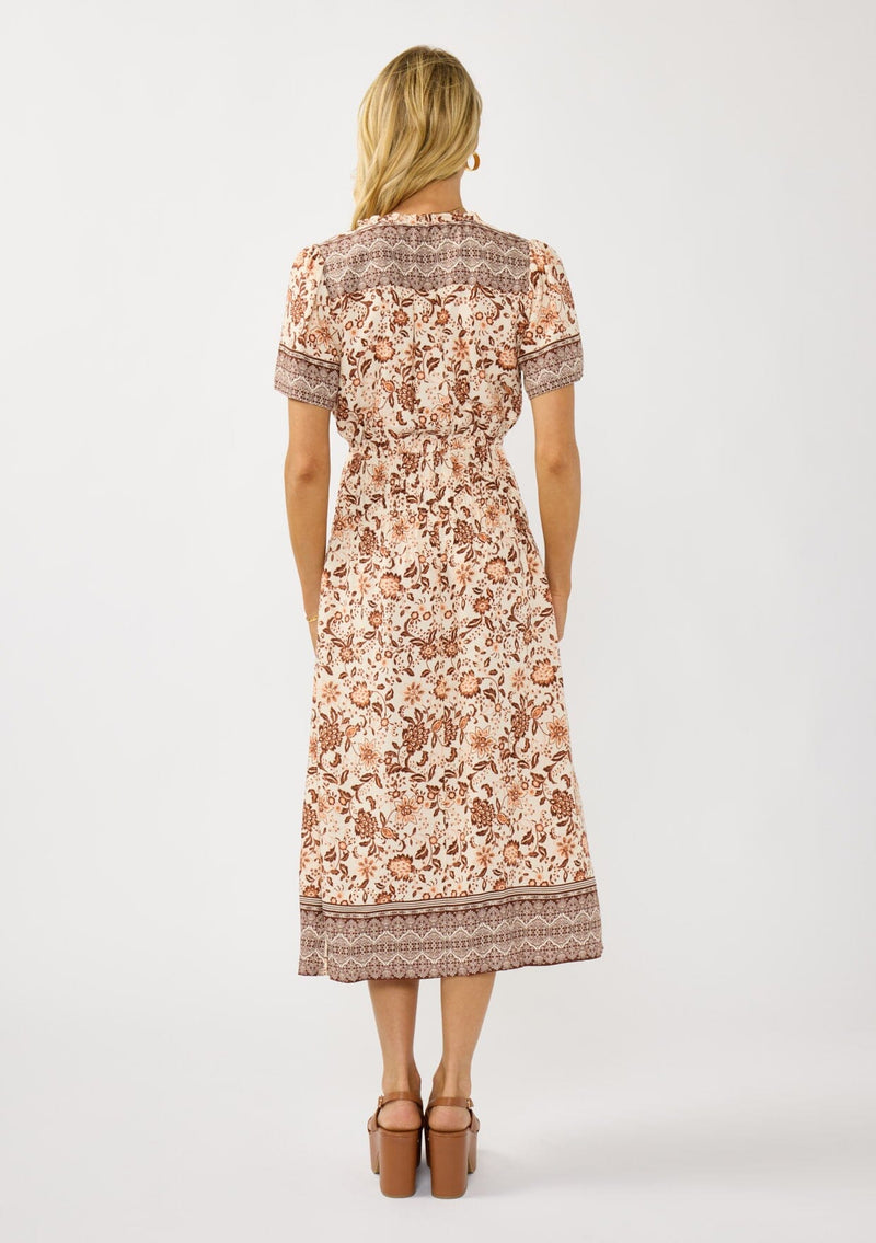 [Color: Natural/Brown] A back facing image of a blonde model wearing a bohemian floral border print mini dress with a split v-neckline, short sleeves, and an elastic waist. 