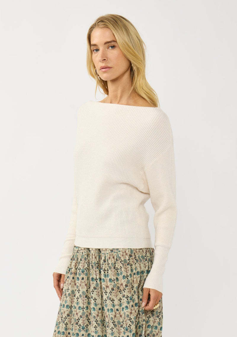 [Color: Natural] A side facing image of a blonde model wearing an off white waffle knit pullover sweater. With long sleeves, a relaxed fit, and a wide neckline that can be worn off the shoulder.