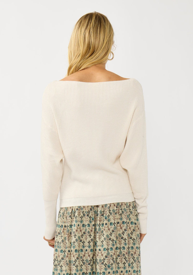 [Color: Natural] A back facing image of a blonde model wearing an off white waffle knit pullover sweater. With long sleeves, a relaxed fit, and a wide neckline that can be worn off the shoulder.