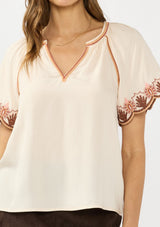 [Color: Sand] A detail image of a brunette model wearing a light beige top with embroidered sleeves. Styled with a brown suede skirt. 