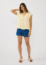 [Color: Yellow/Cream] A front facing image of a brunette model wearing a yellow floral print top with a v-neckline, shirred yoke, and short flutter sleeves. 
