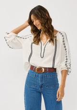 [Color: White/Black] A front facing image of a brunette model wearing a white cotton top with black embroidered details. A classic boho top with a split v-neckline, button front, and long sleeves.  
