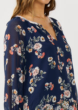 [Color: Navy/Coral] A detail image of a brunette model wearing a navy floral blouse with a split v-neckline, tie detail, and long sleeves with adjustable flutter cuffs. 