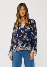 [Color: Navy/Coral] A front facing image of a brunette model wearing a navy floral blouse with a split v-neckline, tie detail, and long sleeves with adjustable flutter cuffs. 