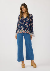 [Color: Navy/Coral] A front facing image of a brunette model wearing a navy floral blouse with a split v-neckline, tie detail, and long sleeves with adjustable flutter cuffs. 