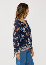 [Color: Navy/Coral] A side facing image of a brunette model wearing a navy floral blouse with a split v-neckline, tie detail, and long sleeves with adjustable flutter cuffs. 