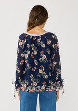 [Color: Navy/Coral] A back facing image of a brunette model wearing a navy floral blouse with a split v-neckline, tie detail, and long sleeves with adjustable flutter cuffs. 