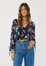[Color: Navy/Coral] A front facing image of a brunette model wearing a navy floral blouse with a split v-neckline, tie detail, and long sleeves with adjustable flutter cuffs. 