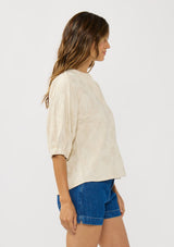 [Color: Natural] A side facing image of a brunette model wearing a beige linen-blend top featuring jacquard detailing, a button-front closure, and elbow-length sleeves with elasticized cuffs.