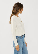 [Color: White] A side facing image of a brunette model wearing a cable knit sweater with a button front. Effortless and easy to style, featuring a classic ribbed trim and crew neckline. A classic year round sweater with a cozy fit. 