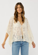 [Color: Natural] A front facing image of a brunette model wearing a beige sheer lace cover up with floral embroidery. Designed with wide kimono sleeves and a delicate tie front detail for easy styling. Perfect for the beach or vacation styling. 