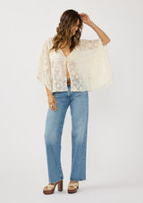 [Color: Natural] A front facing image of a brunette model wearing a beige sheer lace cover up with floral embroidery. Designed with wide kimono sleeves and a delicate tie front detail for easy styling. Perfect for the beach or vacation styling. 