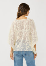 [Color: Natural] A back facing image of a brunette model wearing a beige sheer lace cover up with floral embroidery. Designed with wide kimono sleeves and a delicate tie front detail for easy styling. Perfect for the beach or vacation styling. 