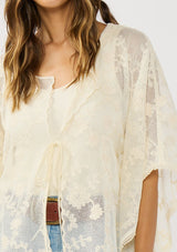 [Color: Natural] A front facing image of a brunette model wearing a beige sheer lace cover up with floral embroidery. Designed with wide kimono sleeves and a delicate tie front detail for easy styling. Perfect for the beach or vacation styling. 