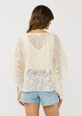 [Color: Natural] A back facing image of a brunette model wearing a beige sheer lace cover up with floral embroidery. Designed with wide kimono sleeves and a delicate tie front detail for easy styling. Perfect for the beach or vacation styling. 
