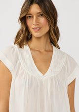 [Color: White] A detail  image of a brunette model wearing a flowy white bohemian top with short sleeves, a v-neckline, raw trim edges, and a shirred yoke. 