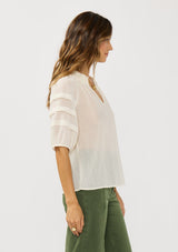 [Color: Ivory] A side facing image of a brunette model wearing an ivory sheer top with a split v-neckline and elbow length sleeves with pleats. A casual warm weather top for resort season. 