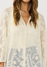 [Color: Natural] A detail image of a brunette model wearing a sheer lace blouse with floral embroidery. Featuring a classic button up design, split v-neckline, and long sleeves. A breezy top with no lining for warm weather styling. 